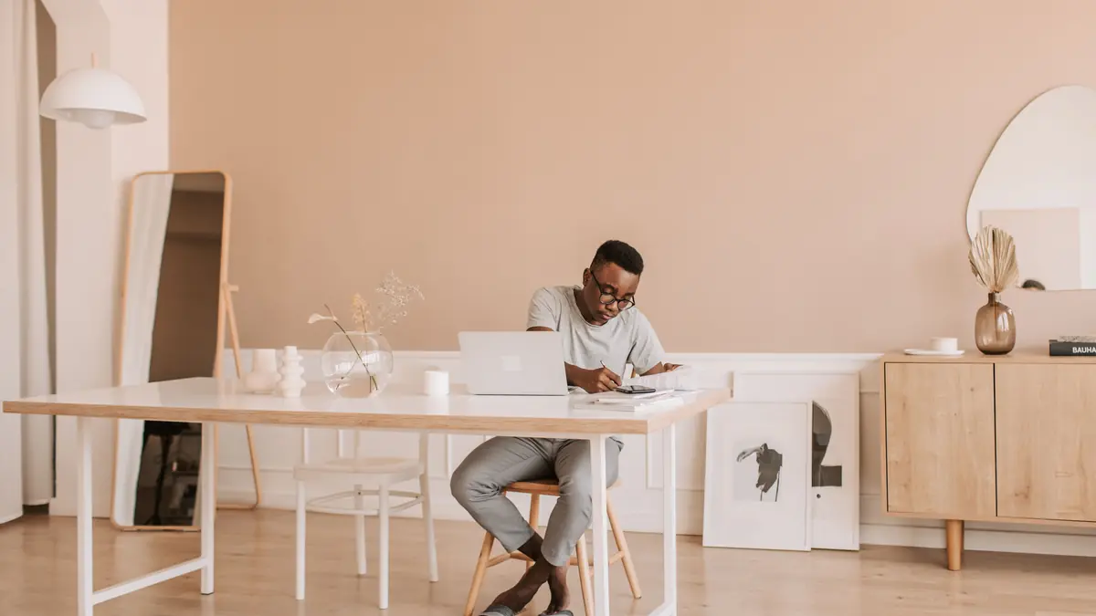 Man writing in a notebook beside a laptop at a white desk in a bright home office
